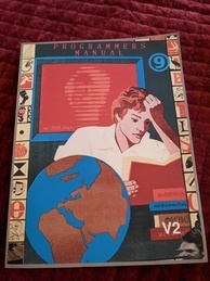 Cover of the 9front Programmer's Manual V2. Features a person intently reading a book while several more books are in a pile waiting to be read and a globe sits next to the reader. A red screen is behind the reader with the text 'up 2920 days' with the rest of the message obscured. The 9front logo is in the upper-right corner, and the indicator V2 is in the bottom-right. 