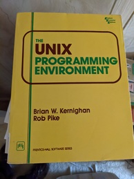 The UNIX Programming Environment book cover. Yellow with the title written in brown and green and circled with a brown border. Has a vertical green and brown line running down the left side